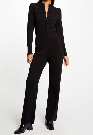 Femme portant un pull côtelé noir zippé, un pantalon noir taille haute à jambes larges et des bottines noires en cuir verni.