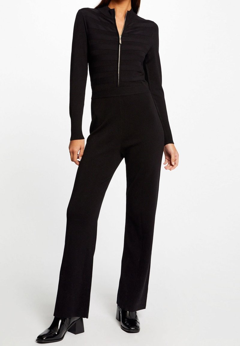 Femme portant un pull côtelé noir zippé, un pantalon noir taille haute à jambes larges et des bottines noires en cuir verni.
