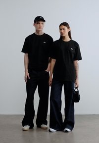 Μαύρα υπερμεγέθη t-shirts, άνετο σκούρο τζιν και μαύρο καπέλο. Σημαντικές λεπτομέρειες περιλαμβάνουν ένα μικρό λογότυπο στα πουκάμισα και μια μαύρη τσάντα χειρός.