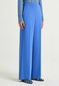 Pantaloni ampi in tessuto blu brillante, caratterizzati da una texture liscia e tasche laterali, abbinati a scarpe blu e nere con motivo.