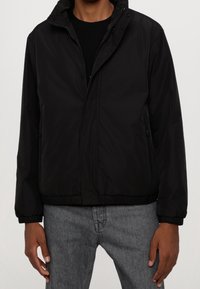 Černá voděodolná bunda s předním zipem, stojáčkovým límcem, elastickými manžetami a bočními kapsami, hladká struktura a volný střih.