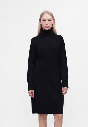 Femme debout portant une robe noire à manches longues et col roulé, longueur genou, sur un fond blanc uni.