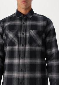 Chemise en flanelle à motifs carreaux noirs et gris. Dispose de deux poches à l'avant, de boutons et d'un logo en étiquette. Fabriquée en tissu texturé doux.
