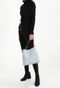 Sac fourre-tout en cuir bleu clair avec des coutures sombres contrastantes et deux poches latérales, présentant un design doux et structuré ainsi qu'une bandoulière réglable.