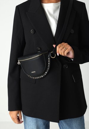 Persona che indossa un blazer doppiopetto nero, una maglietta bianca, jeans blu e tiene in mano una borsa a tracolla nera con borchie, con cerniera dorata e logo MEXX.