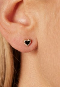 Herzförmiger Steckerohrring mit einem schwarzen Zentrum, umgeben von klaren Strasssteinen, auf goldfarbenem Metall eingesetzt. Nahaufnahme am Ohr.