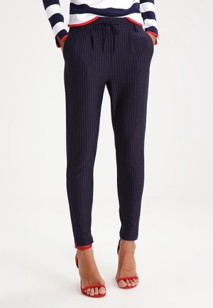 Vrouw draagt marineblauwe broek met witte krijtstrepen, een taille met trekkoord en rode sandalen met hak, handen in de zakken.