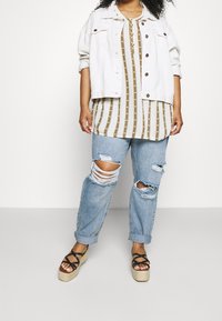 Giacca di jeans bianca sopra una maglietta a righe color sabbia e olivo, abbinata a jeans chiari sdruciti e sandali espadrillas neri con cinturini.