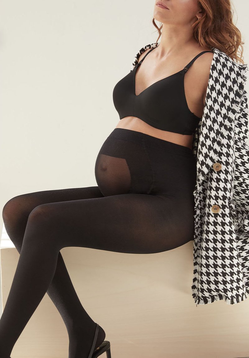 Reggiseno da allattamento nero, collant da gravidanza neri opachi e un cappotto a motivo houndstooth. La modella è seduta su una superficie in legno chiaro.