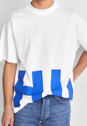 Homme portant un t-shirt blanc surdimensionné avec une grande impression abstraite bleue, partiellement rentré dans un jean en denim bleu, debout devant un fond uni.