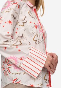 Camicia colorata con bottoni, realizzata in tessuto leggero. Presenta motivi di sole, stelle e occhi in rosa, rosso e marrone. Polsini a righe rosse e bianche.