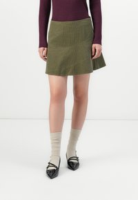 Grön kordflodds A-linje kjol med en texturerad yta, med snedställda sömmar. Matcherade med krämfärgade strumpor och svarta lackskor.