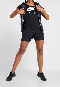 Conjunto deportivo negro que incluye una camiseta de malla de Nike con el logo "AIR", pantalones cortos negros con diseño en capas y zapatillas negras.