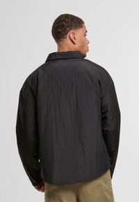 Fekete, gyűrött anyagú ing gallérral és gombos mandzsettákkal, lazán illeszkedő fazonnal és kerekített hemnél, hátulról bemutatva.