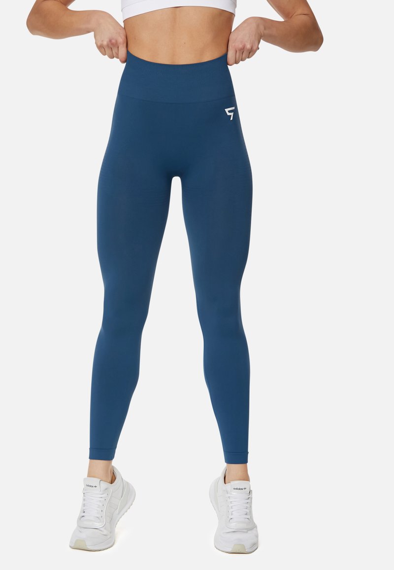 Höga blå leggings gjorda av stretchigt material, med en mjuk yta och en liten logotyp på höften. Kombineras med vita sneakers.