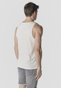 Uomo che indossa una canottiera bianca senza maniche e pantaloncini di jeans grigi arrotolati, in piedi di spalle su uno sfondo semplice.