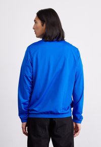 Casaco azul feito de material liso, com gola alta, mangas longas e barra elástica. Usado sobre calças pretas, visto de trás.