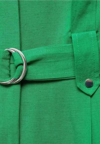 Ceinture en tissu vert avec une boucle en métal argenté et un détail en bouton-pression, présentant une texture lisse et un design structuré.