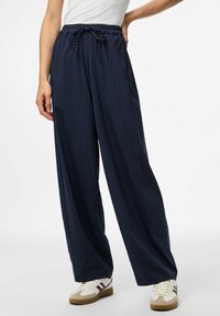 Sieviete, kas ģērbusies tumši zilās svītrainās platās biksēs ar auklu jostasvietā un baltajās kedās ar brūnām akcentu detaļām, stāvot pret baltu fona.