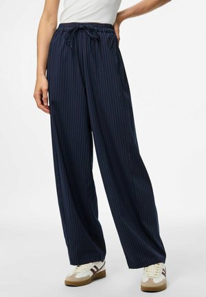Femme portant un pantalon large à rayures verticales bleu marine avec une taille à cordon et des baskets blanches avec des accents bruns, se tenant devant un fond blanc.