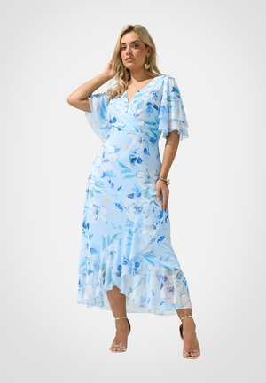 Vrouw draagt een lichtblauwe bloemenprint wikkeljurk met vlindermouwen en een geplooide zoom, poseert met één hand bij haar haar en draagt gouden sandalen.