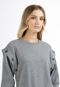 Grijze sweater met lange mouwen, ronde halslijn en decoratieve bronzen knopen op de schouder. Zachte stof, casual pasvorm, minimalistisch ontwerp.