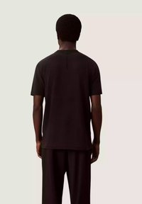 Czarna koszulka z krótkim rękawem o prostym kroju, z okrągłym dekoltem i miękką fakturą, noszona z dopasowanymi czarnymi spodniami, widziana od tyłu.