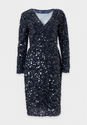 Femme portant une robe noire à manches longues pailletée avec un décolleté en V transparent, debout, une main sur la hanche, chaussée de talons blancs ouverts.