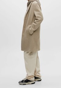 Manteau en laine beige au design structuré avec une fermeture à un seul bouton. Assorti à un pantalon clair et des baskets noires avec des accents gris.