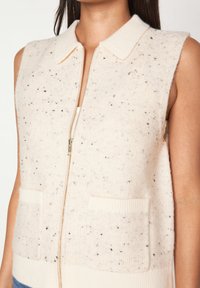 Gilet sans manches de couleur crème avec un col, en tissu en laine texturé agrémenté de taches noires, fermeture éclair à l'avant et deux poches avant.