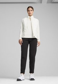 White quilted vest over a white long-sleeve top, paired with black tapered pants and white athletic shoes. Simple design, minimal details.