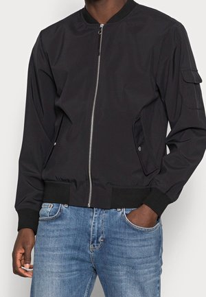 Homme portant une veste bomber noire avec fermeture éclair devant, poches à rabat sur les côtés et sur la manche, associée à un jean bleu, main dans la poche.