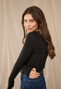 Jauna moteris ilgais rudos spalvos plaukais, vilkinti juodą ilgarankovį viršutinį drabužį ir mėlynas džinsines kelnes, šypsosi ir žiūri per petį priešais smėlio spalvos foną.