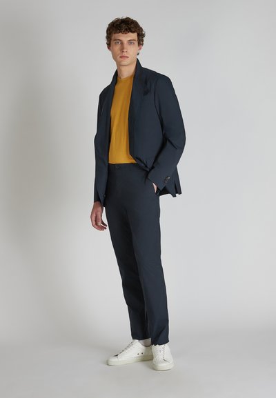Granatowy garnitur o dopasowanym kroju, zestawiony z żółtą koszulką i białymi trampkami. Gładka faktura z delikatnymi detalami na marynarce.