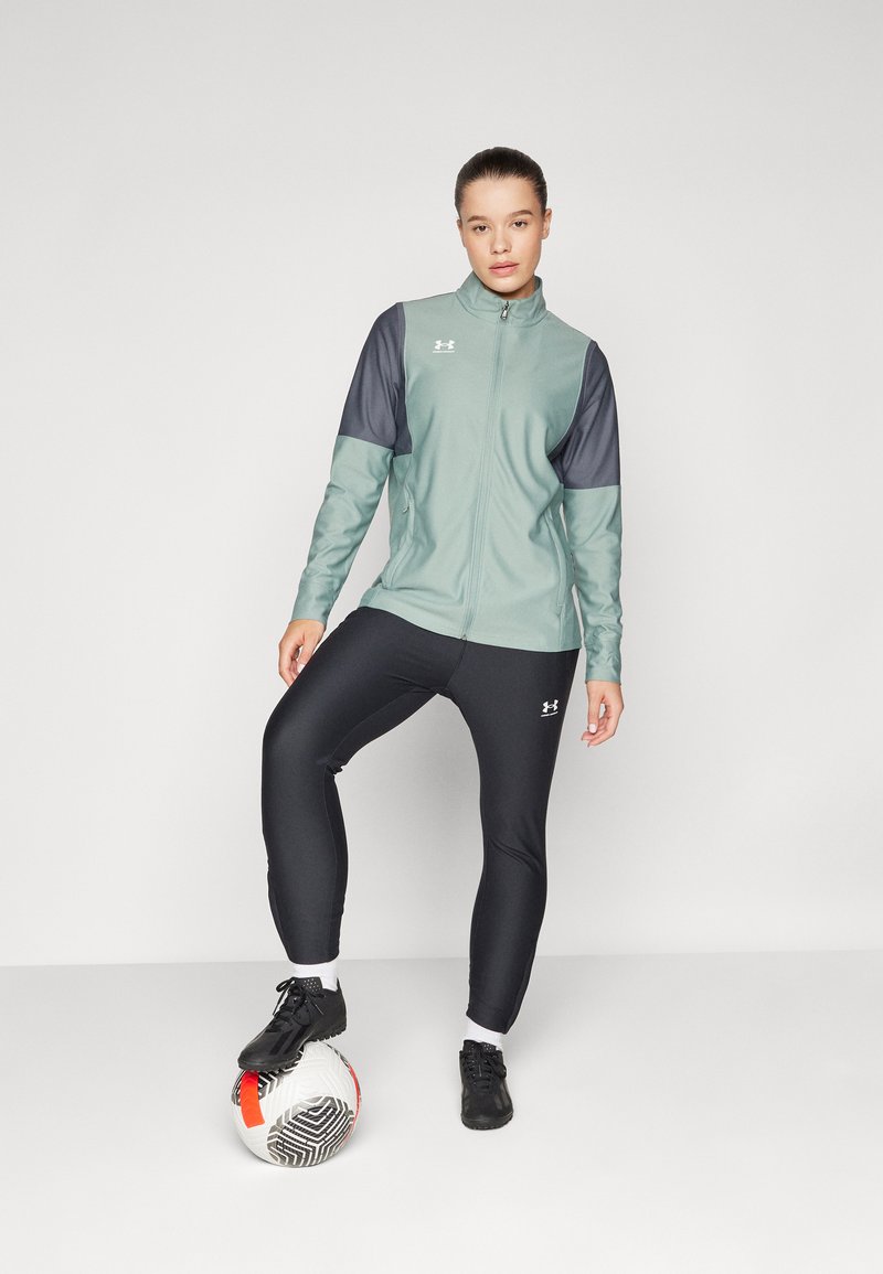 Giacca atletica verde chiaro con accenti grigio scuro, abbinata a leggings neri e scarpe da ginnastica nere, in piedi accanto a un pallone da calcio.