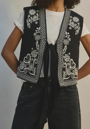 Femme portant un gilet noir avec une broderie florale blanche sur un t-shirt blanc et un pantalon noir, face à l’avant, les mains sur les hanches.