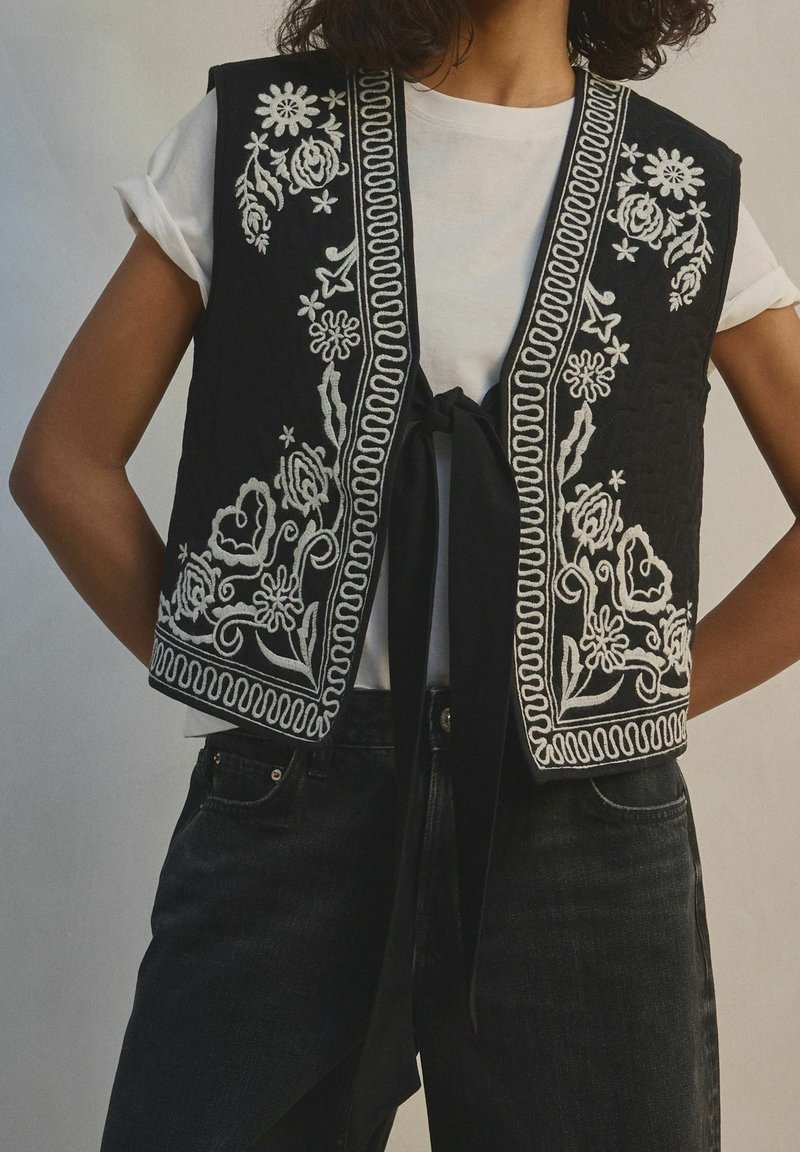 Femme portant un gilet noir avec une broderie florale blanche sur un t-shirt blanc et un pantalon noir, face à l’avant, les mains sur les hanches.