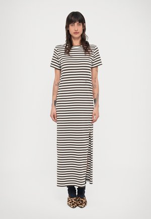 Γυναίκα με σκούρα μαλλιά που φοράει καφέ και λευκό ριγέ μάξι φόρεμα με κοντά μανίκια, μαύρους ζεστά ποδιών και παντόφλες με λεοπάρ σχέδιο, στέκεται όρθια.