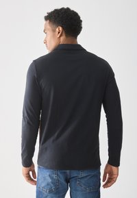Man with short curly hair wearing a black long-sleeve shirt and blue jeans, facing away against a plain light background.
