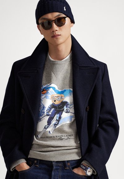 Temno modri volneni plašč čez sivo jopico z grafiko medveda na smučeh, kombinirano s temnimi kavbojkami in sončnimi očali, dopolnjeno z modro uro.