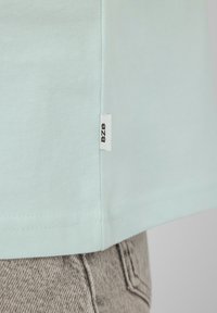 Ourlet en tissu bleu clair avec une petite étiquette blanche marquée « B7B » au-dessus d'une poche de jean gris.