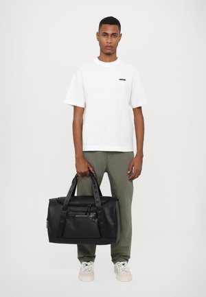 Borsa duffel nera con doppie maniglie, tasca frontale con cerniera e design elegante. Il modello indossa una t-shirt bianca e pantaloni verde oliva.