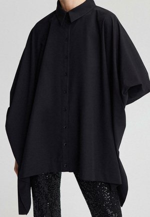 Person iført løs sort skjorte med knapper og brede ærmer samt sorte palietbukser mod en ensfarvet baggrund.