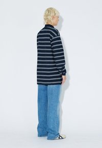 Oversized navy long-sleeve shirt with white stripes, paired with loose blue jeans and beige shoes, view from the back on a plain background.