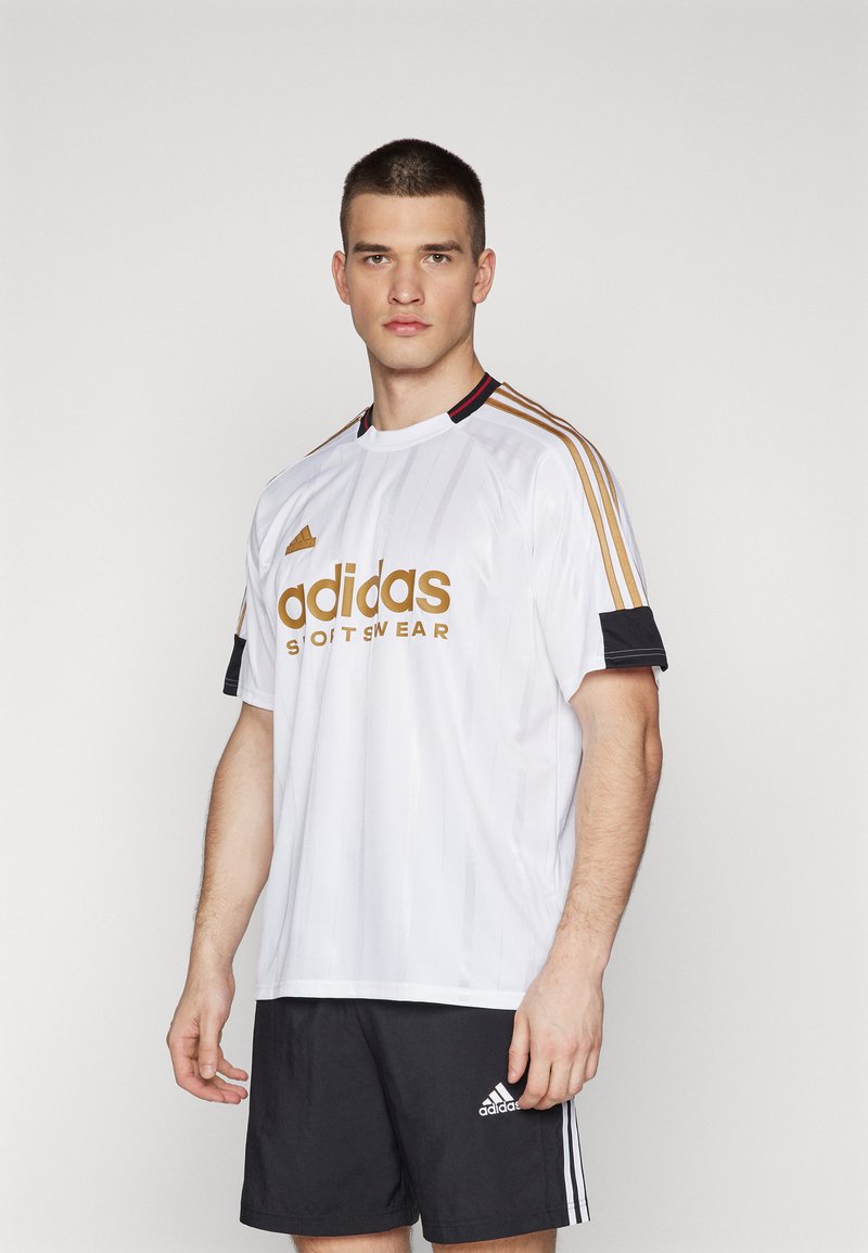 Balta „Adidas“ sportinė maikutė su auksiniu logotipu, juodomis akcentinėmis rankovėmis ir trimis auksinėmis juostelėmis ant pečių, derinama su juodais šortais.