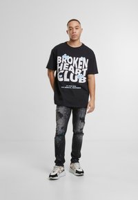 Czarna koszulka z napisem "BROKEN HEART CLUB" na przodzie w dużej białej czcionce, w połączeniu z przetartymi czarnymi dżinsami zdobionymi kolorowymi plamami farby. Białe sneakersy.