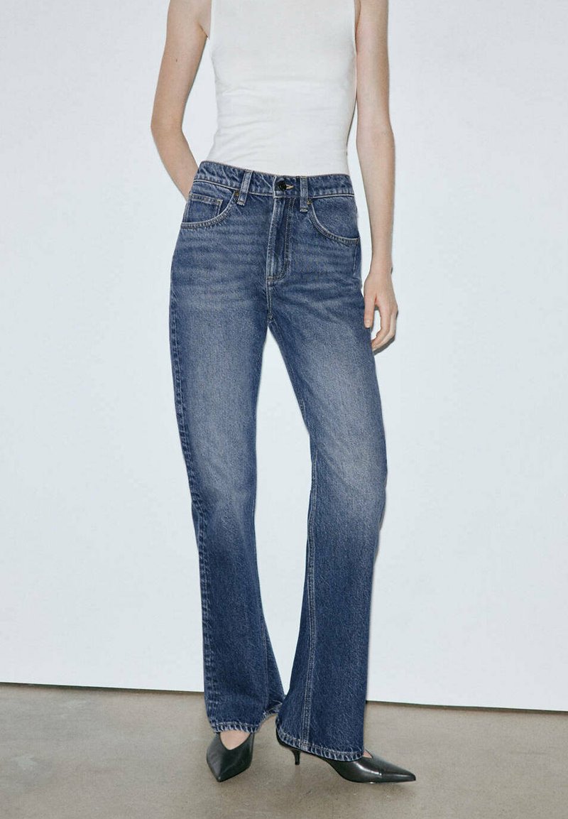 Personne portant un jean bleu taille haute à jambes droites, un top blanc sans manches et des talons noirs pointus, debout devant un fond uni.