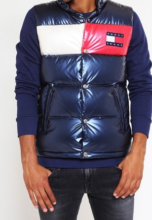 Mann trägt eine glänzende Steppweste in Marineblau, Weiß und Rot mit Tommy Jeans-Logo, darüber ein marineblaues Langarmshirt und dunkle Jeans.