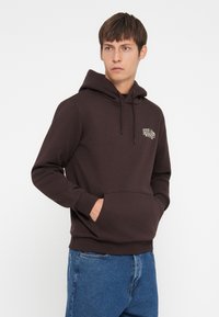 Bruine katoenen hoodie met een kangoeroezak, capuchon met trekkoord en een logo op de borst, gecombineerd met blauwe denim jeans.
