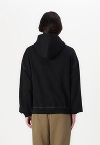 Sweat à capuche noir avec une coupe ample, une grande capuche et des détails de couture blanche. Le tissu semble doux, avec une texture lisse et une silhouette raccourcie.